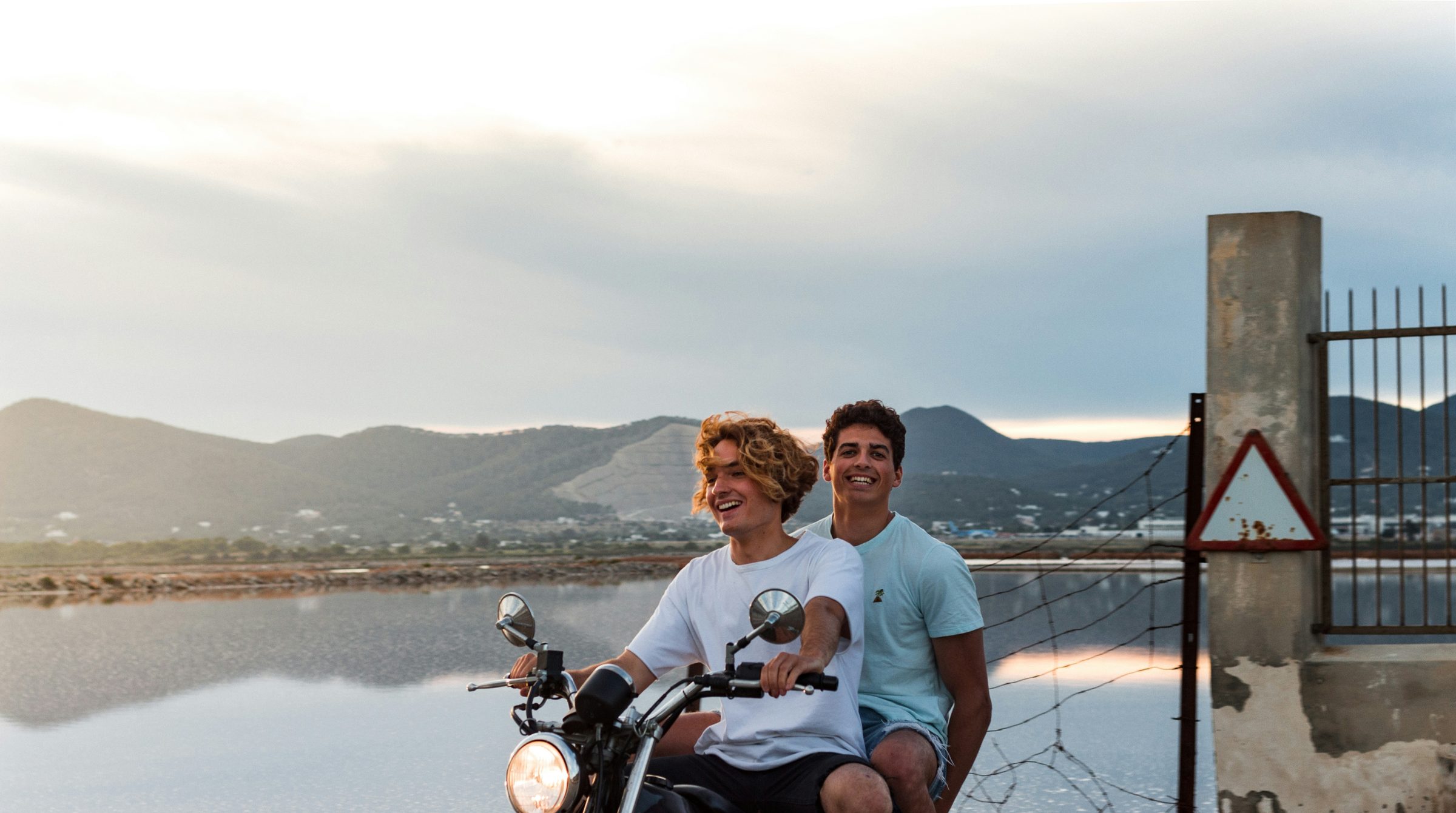 Dos amigos riendo sobre una moto junto al agua
